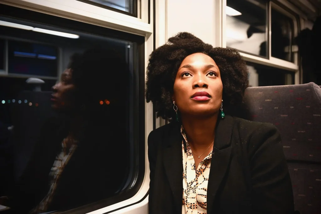 Mulher negra sentada junto à janela de um trem à noite, olhando para cima enquanto seu reflexo aparece no vidro.